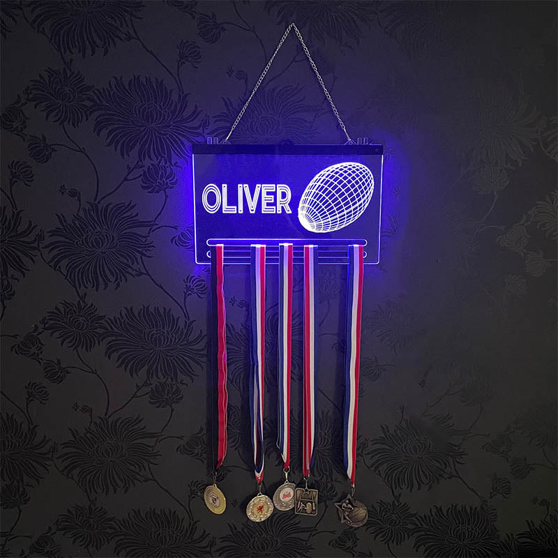 Close-up of a glowing acrylic rugby medal hanger with a name engraved at the top and a detailed rugby ball design, showcasing vibrant blue LED lighting